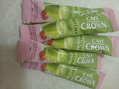 The image displays four individually packaged sachets of strawberry-flavored matcha latte from "Café Crown". The sachets are light pink on the left and transition to a pale green with a white logo and text on the right. Each sachet features an illustration of a glass of matcha latte with latte art and three strawberries. Text on the sachets includes "ÇİLEK AROMALI", "STRAWBERRY FLAVOURED", "MATCHA LATTE", "CAFÉ", and "CROWN". The sachets are arranged in a staggered, overlapping manner on a textured, light-colored surface. The overall scene is a product arrangement, not indicative of any specific location within Balçova, Turkey, or any other city.