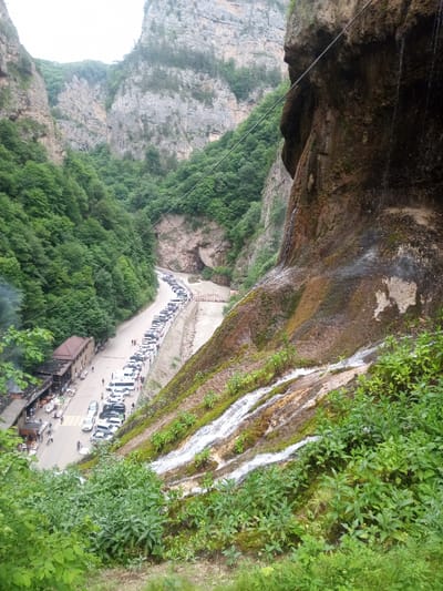 The image depicts a scenic mountain pass with a road winding through a lush green valley. A long line of parked cars, primarily white SUVs and sedans, are visible along the side of the road. People are walking along the road and on the adjacent sidewalk. On the left side of the road, there is a building with an outdoor seating area, possibly a restaurant or cafe, with several tables and umbrellas set up. A small waterfall cascades down a rocky cliff face on the right side of the image, with vegetation growing around it. The mountains are covered in dense green foliage. A thin cable stretches diagonally across the upper right portion of the image. The scene appears to be a popular tourist destination, with many visitors and vehicles present. The location is identified as Khushto-Syrt, Russia.