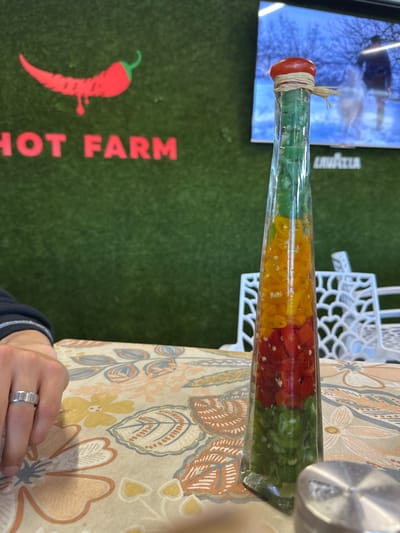 The image shows a long, slender glass bottle filled with layers of green, yellow, and red chopped peppers, sealed with a red cap and tied with twine, resting on a table covered with a floral-patterned tablecloth. A white decorative chair back is visible near the bottle. A sign for "Hot Farm" with a chili pepper logo is visible behind the table. In the background, there is a television screen and a person wearing a ring and a black jacket sleeve. The image was taken in Beloslav, Bulgaria.