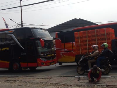 In Megamendung, Indonesia, a dark red and black bus with "JETBUS PARIWISATA" and "BUP-17" written on it is parked on the right side of the road. To its right, a red bus is loaded with orange crates. Two men on a motorcycle, wearing helmets, are passing the buses. One man in a brown jacket is driving, while the other in a black jacket and blue pants is the passenger. In the foreground, a man wearing a red shirt and a yellow vest is crouching on the sidewalk, appearing to be working with a tool. Overhead, power lines crisscross the cloudy sky, and a flag is visible on a pole on the left side of the image.