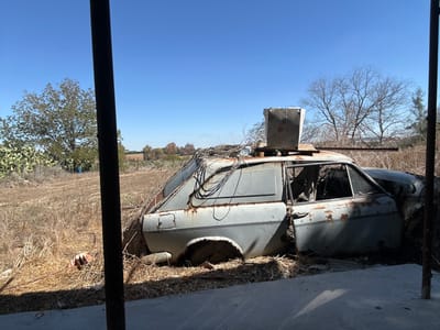 The image depicts a static outdoor scene within the Gderot Regional Council, Israel. The central subject is a derelict, heavily rusted light-colored sedan or hatchback car, resting amidst dry, overgrown vegetation under a clear blue sky. The vehicle is missing its front driver's side wheel, has broken or absent windows, and damaged, partially open doors exposing the interior to debris. A rectangular concrete block is affixed to the car's roof via visible supports, and numerous black electrical wires are draped over its rear and side panels. A long, dark metal pole extends horizontally from the roof area towards the right. The car is situated in a field of dry grasses and weeds. In the immediate foreground, a dark vertical metal post partially obstructs the view on the left, with a concrete surface visible below it. To the left of the vehicle, green cactus plants and a leafy tree are visible. The background features distant agricultural fields and additional trees, some bare, under the bright sky. No people are present.