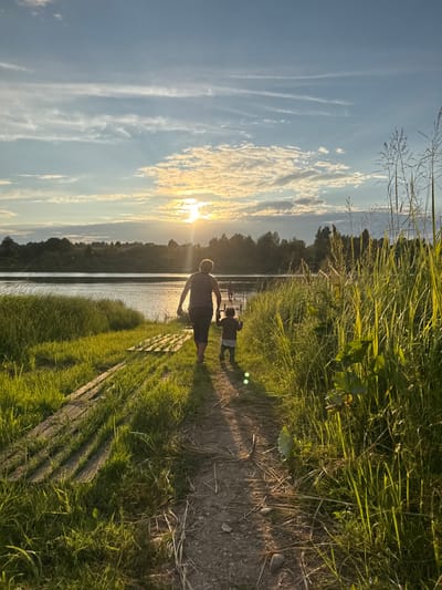 In Sitno, Russia, at sunset, two individuals, an adult and a child, walk away from the camera along a dirt path bordered by tall grass. The adult, wearing a striped tank top and dark pants, is on the left, and the child, in a dark shirt and light pants, is on the right. They are walking towards a body of water, likely a lake or river, with a tree-lined shore in the distance. The sun, low in the sky, casts a warm glow on the scene, creating lens flare and highlighting the clouds. Wooden planks are visible to the left of the path, partially overgrown with grass. The environment is natural and serene, suggesting a rural or park setting.