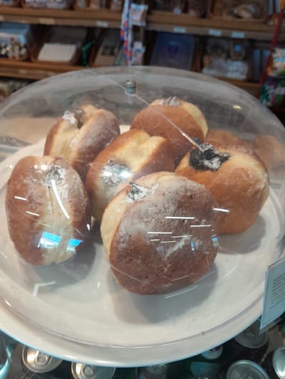 In Cimișlia, Moldova, a transparent dome covers a white plate displaying several pastries dusted with powdered sugar. The pastries appear to be filled, with dark filling visible on some. The scene is set inside a bakery or a similar establishment, as indicated by shelves with various products in the background, including what appear to be bread loaves and other baked goods. A price tag or informational card is visible to the right of the pastries. The lighting suggests an indoor setting, possibly a display case within a store.