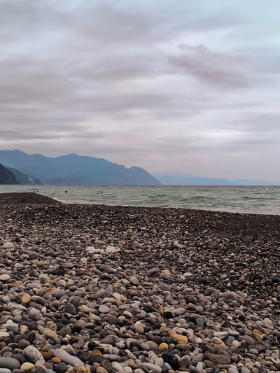 A pebble beach meets the wavy, grey sea under a cloudy sky, with a range of hazy, blue mountains in the distance. The pebbles are predominantly dark grey and brown, with some lighter grey and tan stones mixed in. The water is choppy, with visible waves rolling towards the shore. The mountains in the background are layered, with the closest appearing darker and more defined, while the furthest are lighter and blend into the sky. The sky is overcast with a mix of grey and lighter grey clouds, suggesting a cool or damp day. There are no visible people in the image. The scene is serene and natural, evoking a sense of a secluded coastline.