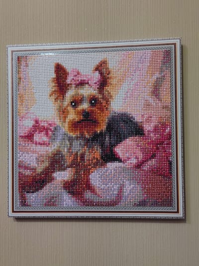 The image shows a framed diamond painting of a Yorkshire Terrier dog. The dog is depicted with a pink bow on its head and a dark collar. It is surrounded by flowers in shades of pink, white, and red. The diamond painting is hanging on a beige, textured wall, likely in a residential setting in the city of Ostafievo, Russia. The lighting suggests it is daytime, and the overall scene is static, with no visible people or actions taking place.