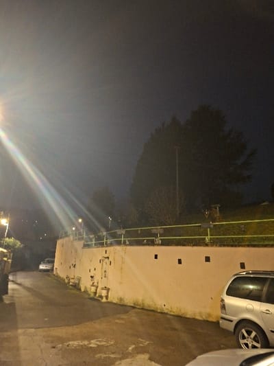 At night, a car is parked beside a high wall with dark squares. A green handrail runs along the wall. The wall has various protrusions and cracks. Beyond the wall, trees are visible against a darkening sky. Light reflects off the street. Other streetlights twinkle in the distance.