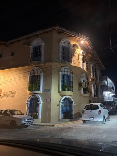 A multistory tan building with arched, grilled windows and blue trim is illuminated at night. A small white car is parked on the sidewalk next to the building, with other cars parked nearby. Power lines crisscross in the sky above. The building's facade features a sign that reads "La Cantera." Street lighting casts shadows, and the sky is dark. The image appears to be taken from inside a vehicle, with part of the car visible.