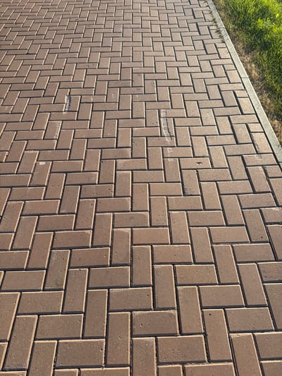 The image shows a close-up, high-angle view of a brown brick paver pathway. The pavers are laid in a herringbone pattern. To the left of the pathway, a strip of green grass is visible, suggesting an outdoor setting such as a park or a garden. The lighting indicates it is daytime, with shadows cast by the pavers. There are some minor imperfections on a few of the pavers, such as chips and scuffs. No people, animals, or text are visible in the image. The overall scene depicts a section of an outdoor pathway.