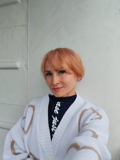 A middle-aged woman with short, peachy-blonde hair and light skin is looking towards the camera with a slight smile. She is wearing a dark blue t-shirt with white cursive writing and a white cardigan with tan heart patterns. She has on subtle makeup, including eyeliner and lipstick. She is also wearing a small, sparkly heart-shaped earring in her left ear. The woman is positioned in front of a light-colored, textured wall with some vertical seams. The overall lighting is soft and natural. The scene appears to be outdoors in an urban setting, possibly in Vytegra, Russia.