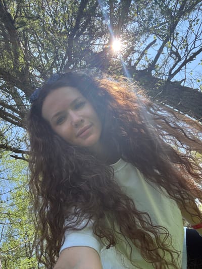 A woman with long, curly brown hair is pictured from a low angle, looking up at the camera. She is outdoors, with a tree canopy and blue sky visible above. Sunlight flares into the lens from the upper left of the frame. She is wearing a light-colored shirt. The background suggests a natural, possibly rural or park setting. No specific location within Bichiktu-Boom, Russia, can be determined from the visual information.