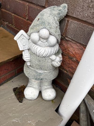 An outdoor close-up depicts a grey garden gnome figurine. The gnome features a textured, fuzzy surface on its pointed hat and body, resembling flocking or a similar material. Its prominent nose, full beard, hands, and feet are sculpted from a smoother, stone-like material, showing some speckled discoloration. The gnome holds a small, house-shaped sign in its hands, but no text is legible on it.
The figurine rests on a light grey concrete or paved surface. A patch of dark green and brown moss grows on the concrete near the gnome's feet. To the left and upper right, a reddish-brown brick wall provides a backdrop. A white PVC pipe runs diagonally across the top right of the frame, resting against the brickwork. Small water droplets are visible on the pipe and the damp paved surface, indicating recent precipitation. The scene captures a static arrangement within an outdoor setting, likely a garden or side of a building in Minsterley, United Kingdom. No specific activity is occurring, and there is no discernible text.