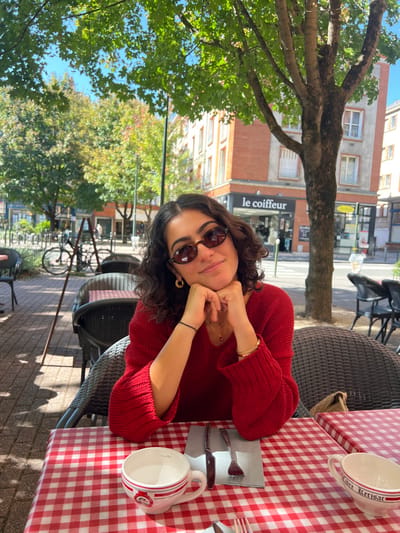 A young female with dark, wavy hair and oval-shaped brown sunglasses is seated at an outdoor café table, smiling towards the viewer. She is wearing a red knitted v-neck sweater, gold hoop earrings, a delicate gold necklace, and a gold-colored beaded bracelet on her right wrist, with a black elastic band on her left wrist. Her hands are clasped under her chin, resting on her forearms.
The table is covered with a red and white checkered tablecloth. On the table, to her left, is a white mug with red and black details, marked "2cl" and partially legible text "Orléans." To her right, a second white mug reads "Libre Kerisac." Between the mugs, a knife and fork are placed on a grey napkin. Dark rattan-style chairs with striped cushions are visible at her table and surrounding tables.
The scene takes place on a paved street or square under natural sunlight, with dappled shadows cast by overhead tree foliage. In the background, a multi-story building with reddish-brown brick upper floors and white window frames is prominent. The ground floor of this building houses a storefront with a sign that reads "Le Coiffeur" in white text on a dark background. A yellow sign is visible to the right of "Le Coiffeur." Further left, several bicycles are parked along the street. The location is **110 Rue de Bourgogne, Orléans, France**, identified by the specific architecture and "Le Coiffeur" sign, which corresponds to "Coiffeur le Coin" at this address.