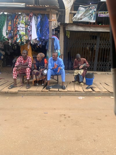 This image depicts a street scene in Egbedi, Nigeria, focusing on a raised wooden platform along a commercial thoroughfare. The foreground consists of a dirt road with scattered small debris.
On the wooden platform, four individuals are seated on low stools. From left to right:
1. A male, dressed in a red and white patterned traditional tunic and trousers, sits with arms crossed, observing. He wears black sandals.
2. A female, wearing a headscarf, a brown and orange patterned dress, and an orange long-sleeved top, gestures with her right hand while holding a small white disc-like object in her left, appearing to communicate. She wears white sandals.
3. A male, dressed in a light blue traditional tunic and trousers, sits with hands slightly open, looking forward. He wears black loafers.
4. A male, wearing a dark red patterned traditional tunic and trousers, sits with a blue cloth draped over his lap, actively shining a black shoe held in his hands. He wears light brown slides. A blue plastic basket containing a dark object is beside him, and another black shoe rests on the platform. A black rectangular mat or kit is positioned on the platform in front of the blue-clad man.
In the background, to the left, a retail stall displays numerous patterned garments hanging for sale beneath a corrugated metal roof topped with several automobile tires. To the right, a shop entrance is secured by a closed metal accordion gate. Above this shop, a banner reads "HERO GLAD ART SIGNS."
