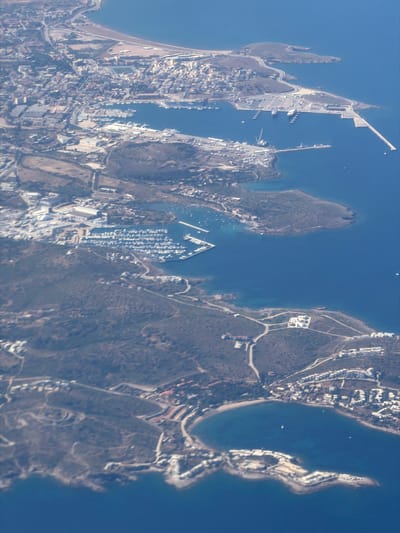 This aerial image shows a coastal area with multiple peninsulas and islands. On the left side, there is a built-up peninsula with a developed harbor featuring docks and infrastructure. Adjacent to this, another peninsula extends into the water, containing a marina with numerous boats. Further to the right, a larger landmass exhibits sparse vegetation and some residential development along the coast. The water between the landmasses is deep blue, suggesting a navigable waterway. The image provides a clear view of the geographical layout and human development in this coastal environment. The location is within the city of Paiania Municipal Unit, Greece.