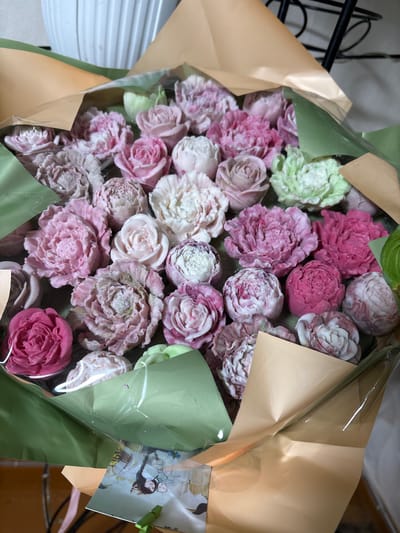 A bouquet of decorative flowers is presented in a display of brown and green wrapping paper. The flowers themselves are crafted from a light-colored material and are shaped like roses, peonies, and other floral varieties. They are colored in various shades of pink, white, and light green. The bouquet is wrapped in clear plastic. A small, folded card is attached to the front of the bouquet, showing a photo of a woman and a baby. The background is out of focus, but suggests an indoor setting with a white, textured object and part of a black metal structure. The image does not provide enough information to determine the specific location within Siberia, Russia, or to describe any actions or interactions of people.