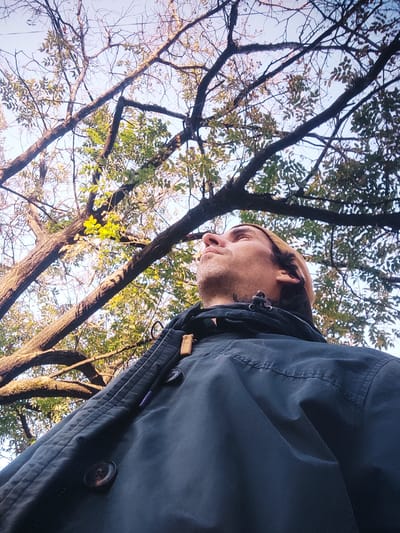 A low-angle, close-up shot captures a man looking upwards through the branches of a tree, which are silhouetted against a pale blue sky. The man, whose face is partially obscured by his chin and the angle of the camera, is wearing a dark jacket and a light-colored beanie. The tree has rough bark and a mix of bare branches and sparse green leaves, suggesting it might be autumn. The scene is likely outdoors, possibly in a park or a residential area with mature trees, such as those found in the city of Remedios de Escalada, Argentina. The lighting is natural, with the sky appearing bright and the trees creating a natural canopy. The image has a slightly grainy texture, and the colors are muted, with a focus on the textures of the bark, leaves, and fabric.