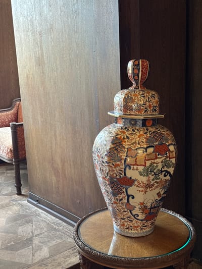 A decorated, antique ceramic vase sits atop a round table with a metallic rim. The vase has a lid and handle, and is covered in intricate floral and geometric patterns. The table and vase are positioned near a light-colored wooden surface and a dark wooden wall. Part of a chair with patterned upholstery is visible in the lower left. The floor has a herringbone pattern. The location is likely within the Reichsburg Cochem, Germany, given the style of the furniture and decor. No people are visible.