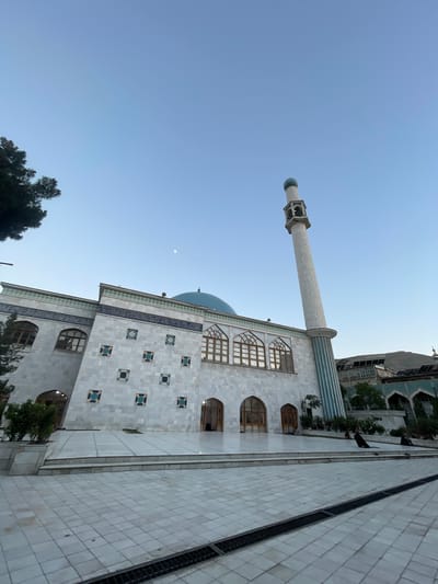 The image shows the exterior of the Blue Mosque in Kabul, Afghanistan. The mosque is a large, white marble building with a blue dome and a tall, slender minaret. The facade of the mosque is decorated with intricate blue and white tilework. In front of the mosque is a large courtyard paved with white tiles. Several people are seated in the courtyard, some facing the mosque. The sky is clear and blue, with a faint moon visible. The overall scene is peaceful and serene.