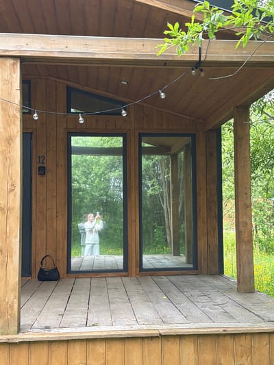 The image displays the entrance area of a modern wooden cabin or building, likely a vacation rental, oriented vertically with the top of the structure to the left. The building features light brown wooden siding and robust wooden beams, suggesting a sturdy construction. Large, rectangular windows are prominent, reflecting the lush green foliage and trees of the surrounding natural environment, along with patches of yellow flowers, possibly a field of dandelions, in the distance. A smaller, dark-framed triangular window is visible on the left side of the structure.

A string of clear globe lights hangs diagonally across the porch area, adding a decorative touch. In the reflection of the lower main window, a person, presumably the photographer, is visible holding a smartphone, capturing the scene. They appear to be wearing light-colored attire.

On the wooden wall to the right of the lower window, the number "12" is prominently displayed, indicating a unit or address number. Below this, a black electronic keypad or lock is mounted next to a dark-framed door. A black handbag with sunglasses resting on it is placed on the wooden floor/porch, suggesting recent arrival or imminent departure. The scene is brightly lit, indicating daytime conditions. The overall impression is that of a tranquil, nature-surrounded retreat in Vozhoiskii, Russia.
