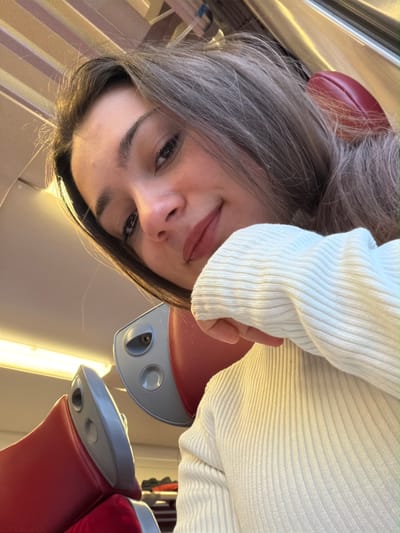 A young female is depicted in a close-up, upward-angled shot, likely a selfie, within the interior of a public transport vehicle, identified as being in Obermodern-Zutzendorf, France. The subject, positioned centrally, has dark hair, dark eyes, and wears a cream-colored, ribbed long-sleeved top. She presents a subtle smile with slightly parted lips and looks directly at the camera. Her left arm, covered by the sleeve, is partially visible. The background features the interior of a train or bus, characterized by red upholstered seats with gray plastic frames. Two seatbacks are partially visible; one is directly behind the subject's head, and the top of another is to the lower left. The vehicle's ceiling is light-colored with horizontal panels, and internal lighting is present. A reflective or metallic strip, possibly part of a window frame or luggage rack, is visible in the upper right quadrant.
