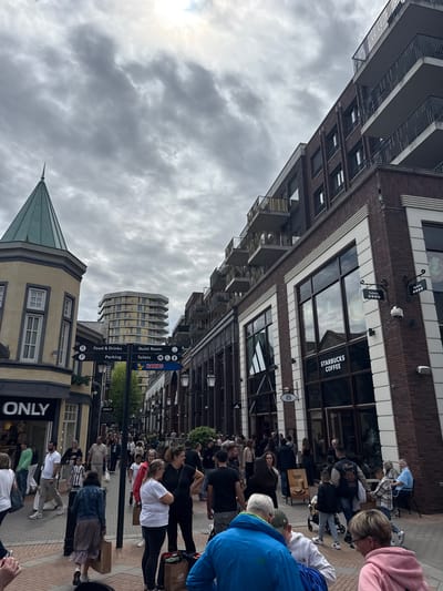 This image depicts a crowded outdoor shopping street within the Designer Outlet Roermond, Netherlands, under an overcast sky. The primary light source is diffused from above, suggesting a cloudy day.

On the left side of the street, a multi-story building with a light-colored facade features a prominent green-spired turret on its corner. Multiple rectangular windows with white frames are visible on this building. The ground floor houses a store with "ONLY" displayed prominently on its signage. Further down the street, additional buildings with similar architecture are visible.

In the center, a black directional sign post extends upwards, featuring multiple white arrows on black panels. These signs indicate "Food & Drinks," "Quiet Room," "Parking," and "Toilets" (accompanied by universal access symbols). Below these, a blue rectangular sign with "HARIBO" in white text points to the right. In the background, a distinctive multi-story building with a light-colored, undulating facade and numerous balconies rises above the other structures.

The right side of the street is lined with multi-story brick buildings featuring dark-framed windows and balconies with dark railings on the upper floors. White architectural trim highlights corners and window surrounds. The ground floor of one of these buildings houses a "STARBUCKS COFFEE" store, identifiable by its large glass windows and prominent sign. Reflections in these windows show parts of the opposite buildings and the sky. Another store with a faint logo (possibly Adidas) is adjacent to Starbucks. Security cameras are mounted on the exterior walls of these buildings.

The pedestrian street is paved with light-colored bricks arranged in various patterns. Numerous individuals, dressed in casual attire, are populating the street. They are seen walking, standing, and interacting. Several people carry shopping bags, including a brown "New Balance" bag visible in the lower right foreground. Some individuals are seated at outdoor tables near the Starbucks entrance. A person in a blue jacket is prominent in the lower left foreground, facing away from the camera. A child in a black and white striped shirt is visible in the middle ground, and a person is pushing a baby stroller near Starbucks. Black bollards and decorative black street lamps are spaced along the pedestrian pathway.