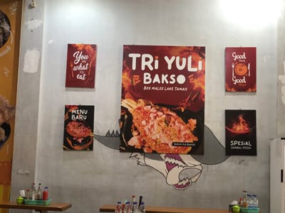 The image depicts a wall in an Indonesian restaurant, likely in Uteungeulinggang. The wall is adorned with several framed posters. On the left, a red poster with the text "You are what you eat" is visible. Next to it, a large central poster advertises "Tri Yul Bakso," with the subtext "Bek Malee Lake Tamah," featuring an appetizing image of bakso. To the right of the central poster, a smaller red poster displays "Good Food is Good Mood." Below the central poster, a dark poster announces "MENU BARU" (New Menu) with an image of bakso. Further to the right, another dark poster showcases "SPESIAL SAMBAL PEDAS" (Special Spicy Chili Sauce) with a fiery chili pepper graphic. The wall itself is painted a light gray and shows some signs of wear. Below the posters, a cartoonish illustration of a shark or monster with sharp teeth is painted on the wall. In the foreground, a wooden table holds a green plastic basket containing various bottles of condiments and sauces, along with napkins. The lighting suggests an indoor environment, likely during the day.
