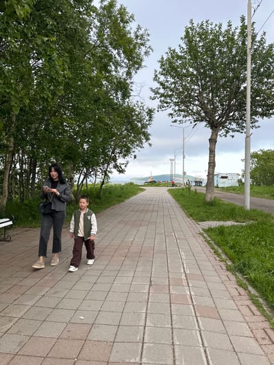 The image depicts a paved walkway in Vilyuchinsk, Russia, bordered by grass and trees. A woman, dressed in a grey jacket and dark pants, walks with a young boy wearing a green and white jacket and brown pants. The woman is looking at her phone, while the boy walks ahead of her. In the background, a body of water can be seen with a boat and some buildings along the shore. The sky is overcast.