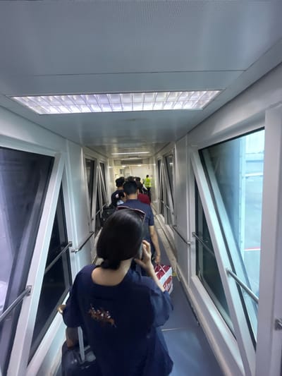 A group of people are walking through an airport jet bridge. The jet bridge is enclosed with large windows on the right side, offering a view of the tarmac and an airplane. The interior of the jet bridge is lit by fluorescent lights in the ceiling. The people are dressed casually and are carrying luggage, suggesting they are boarding or deplaning an aircraft. One person in the foreground is facing away from the camera, looking down at their phone. In the background, a person in a high-visibility vest is visible, possibly an airport worker. The overall scene depicts a common airport transit environment.