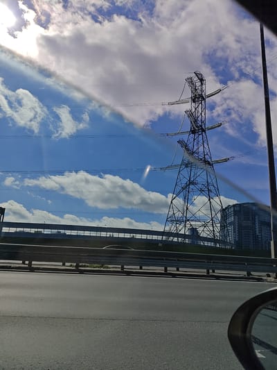 The image depicts a street-level view with a large electrical pylon as the dominant feature against a bright, partly cloudy sky. The pylon is a lattice-steel structure with multiple cross-arms supporting electrical cables. In the background, a modern, multi-story building is visible, suggesting an urban or suburban setting. A highway guardrail runs horizontally across the middle ground, separating the road from an elevated area with some greenery. The foreground shows a paved road, likely a highway, with the edge of a car's side mirror visible in the lower right corner. The lighting suggests it is daytime, with the sun casting bright rays through the clouds, creating lens flares and highlighting atmospheric haze. The overall scene conveys a sense of infrastructure and urban development within a bright, somewhat hazy daylight. The location is identified as Tarasovka, Russia.