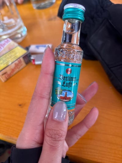 A close-up image depicts a human hand holding a clear, miniature glass bottle of "Berliner Luft" peppermint liqueur. The hand has light skin and fingernails painted with a translucent, light purple/grey polish. The bottle features a teal label with the brand name "Berliner Luft" in stylized white text and an oval illustration of a red-brick building with a prominent tower, likely the Rotes Rathaus in Berlin. The bottle has a silver screw cap with a teal band beneath it. In the blurred background, situated on an orange-brown wooden table, a clear glass partially filled with liquid is visible on the left. To the right of the glass, a yellow and brown "Chesterfield" cigarette pack lies near at least two lighters, one appearing white and another metallic/red. A dark black fabric, possibly a jacket or glove, is present in the upper right background. The scene is located in St. Christoph, Austria.