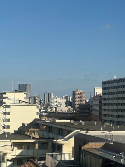 A daytime aerial view reveals a dense urban landscape in Nakano, Japan, under a clear, light blue sky with sparse, high-altitude clouds. The scene is static, devoid of people or visible activity.

In the foreground, several rooftops are visible. To the bottom right, a building section displays a dark brick-patterned facade and a flat, grey roof equipped with multiple white cylindrical vents and a ladder leading to the roof edge. Adjacent to this, another building features light-colored balconies with metal railings, one balcony exhibiting a distinct curved design. A small section of a reddish-brown tiled roof is partially visible at the bottom left.

The midground is dominated by a variety of multi-story buildings. A prominent light-cream colored building on the left features external staircases with white railings on its facade and multiple balconies. To the right, a tall, light-grey building with numerous rectangular windows forms a grid-like pattern. Between these structures, a compacted arrangement of residential and commercial buildings of varying heights and architectural styles, primarily in off-white, beige, and light grey hues, extends into the distance. Small clusters of dormant trees with bare branches are interspersed throughout the urban fabric.

The background showcases the extended cityscape, with numerous high-rise buildings and skyscrapers, including a taller, dark-grey skyscraper visible on the far left, protruding above the surrounding urban structures against the horizon.