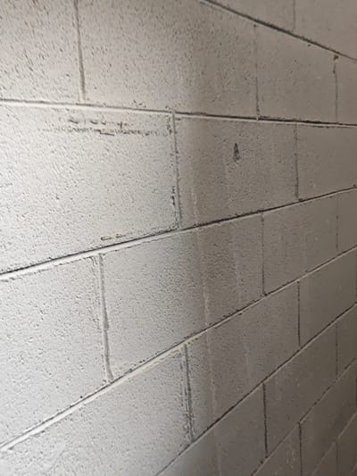 A close-up, low-angle shot captures a textured, gray painted cinder block wall in Settimo Torinese, Italy. The perspective emphasizes the horizontal and vertical mortar lines between the blocks, which recede into the background, suggesting the wall continues out of frame. The surface of the blocks is rough and uneven, with small imperfections and some darker marks visible, indicating age or wear. The lighting appears to be artificial and casts subtle shadows, enhancing the three-dimensional quality of the textured surface. No people or specific objects are discernible on the wall itself.