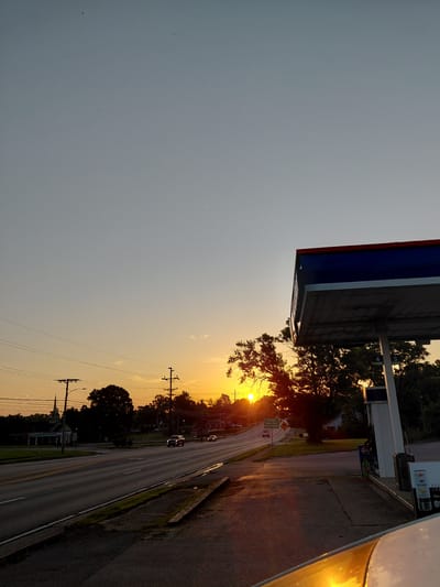 The image depicts a sunset scene at a gas station in Frankfort, United States of America. The sun is low on the horizon, casting a warm glow and long shadows. The sky is a gradient of pale blue to orange. A gas station canopy, with blue and red trim, is prominently featured in the foreground, partially obscuring the view. To the left of the canopy, a road with traffic is visible, with cars driving and a church steeple in the distance. Trees and power lines line the road. The ground in front of the gas station pumps is paved, and a portion of a white vehicle is visible in the lower right corner.