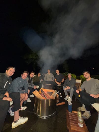 The image depicts nine adult males gathered around a stainless steel fire pit on an outdoor patio at night in Huasca de Ocampo, Mexico. The subjects are arranged in a semi-circle on various outdoor seating, including wooden chairs with light cushions, on a dark paved surface. The central fire pit contains actively burning logs, producing a prominent plume of smoke that rises into the dark, unlit background of obscured foliage. All individuals are casually dressed, predominantly in long-sleeved shirts or jackets, and most are smiling while looking towards the viewer. Specific details include: a male on the far left wearing a backward baseball cap; a male, fourth from the left, displaying a peace sign with his left hand; a male to the right of the fire pit holding a dark beverage bottle; and a male on the far right holding a brown beverage bottle with a yellow label. A low, wooden-slatted table in the lower right foreground holds a blue insulated tumbler and a clear glass containing a liquid. The scene indicates a social gathering.