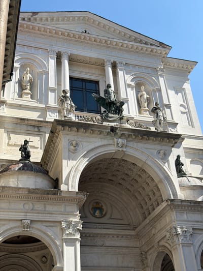 The image shows a detailed view of the facade of the Church of San Lorenzo Martire in Mozzo, Italy. The church's white stone exterior is adorned with classical architectural elements, including columns, pediments, and statues. At the center of the facade is a large arched entrance, above which sits a sculptural group featuring a winged figure. Niches along the upper facade house statues of various figures, likely saints or religious icons. The clear blue sky suggests it is daytime. The building exhibits intricate carvings and decorative friezes.