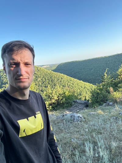 A man with short brown hair and a light stubble is taking a selfie in a mountainous, forested area. He is wearing a black long-sleeved shirt with a large yellow smiley face graphic on the left chest. The man is positioned in the foreground, with his face slightly turned towards the camera and a neutral expression. Behind him, a vast expanse of green, tree-covered hills stretches into the distance. The sky is a clear, pale blue. The immediate foreground consists of dry grass, rocks, and some sparse vegetation. The location is identified as Pryvorottia Druhe, Ukraine.