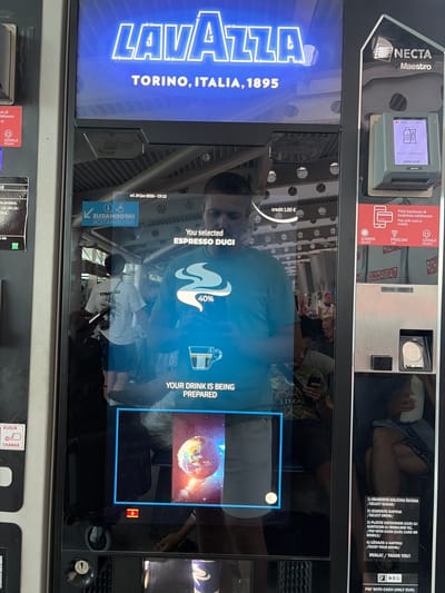The image shows a close-up view of a Lavazza coffee vending machine, likely located in Golubovci, Montenegro. The machine features a large, illuminated "LAVAZZA TORINO, ITALIA, 1895" logo. The touchscreen display is active, showing that an "ESPRESSO DUGI" has been selected and is currently being prepared, with a progress indicator at 40%. A celestial image of Earth is displayed on the screen.

Above the main display, there's a smaller Necta Maestro interface, showing payment options like VISA and contactless payment symbols. Instructions in Croatian and English are visible, detailing how to select sugar, choose a drink, make a payment (cash, card, or mobile), and enjoy the drink. The time "sri. 24 jun 2026-19:12" is displayed, suggesting it's late afternoon or early evening.

Reflections on the glass display reveal people in the background, suggesting this is a public indoor setting, possibly a transportation hub or a shopping area. There are also various labels and instructions on the machine, including currency symbols (EUR) and payment methods. The overall scene conveys a modern, automated beverage service in operation.