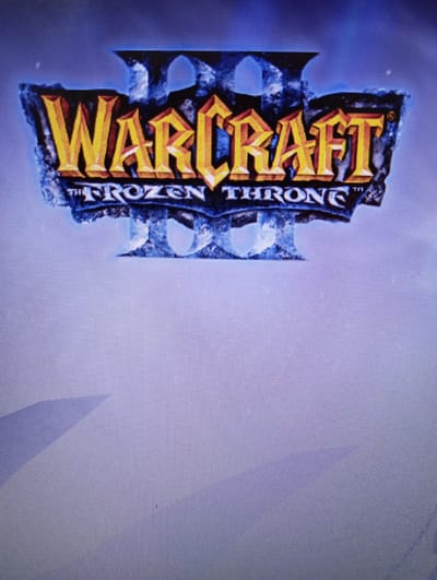 The media file displays the title screen for the game "Warcraft III: The Frozen Throne." The logo is depicted vertically, with "Warcraft" in large, golden, three-dimensional letters at the top, and "The Frozen Throne" written vertically in smaller, white letters to the right of "Warcraft." The background is a gradient of pale blue and white, suggesting a cold, possibly snowy or icy, setting. The "Warcraft" logo itself appears to be carved into or made of stone and ice, with dark blue and grey textures and sharp edges. There are no people visible in the image, only the game's title and background elements. The overall impression is one of a fantasy or epic theme associated with a video game.