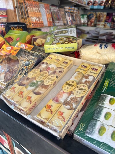 The image shows a display of various sweets and confectionery items in a shop in Dahab, Egypt. On the right side are boxes of "El Shanawany Nougat with Candied Fruits & Peanuts," stacked in a cardboard tray. At the top right, a green box of "Pistachio Lokum Delight" can be seen. To the left, there's a green box labeled "Bavero Lokum Delight." The background reveals more boxes of sweets and an open bag filled with a cotton candy-like substance, labeled "حلوى ان."