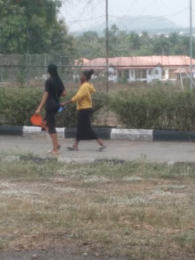 This image, set in Osu, Nigeria, depicts two females walking from left to right on a paved surface adjacent to a curb. The individual on the left wears a black long-sleeved top and a black long skirt, holding an orange paddle-like object in her right hand and a small blue object in her left. Her hair is dark and long. The individual on the right, slightly ahead, wears a yellow hooded sweatshirt and a dark long skirt or trousers, with her hair pulled back. She extends her right arm towards the first female's left hand. Both wear open-toed footwear. A concrete curb, painted with black and white stripes, separates the paved area from a strip of dry, patchy grass in the immediate foreground. Beyond the pedestrians, a row of green bushes fronts a chain-link fence topped with barbed wire. In the background, multiple white buildings with prominent terracotta roofs are situated amidst dense tropical foliage, primarily palm trees. Hazy hills are visible on the distant horizon under a diffuse daylight sky.