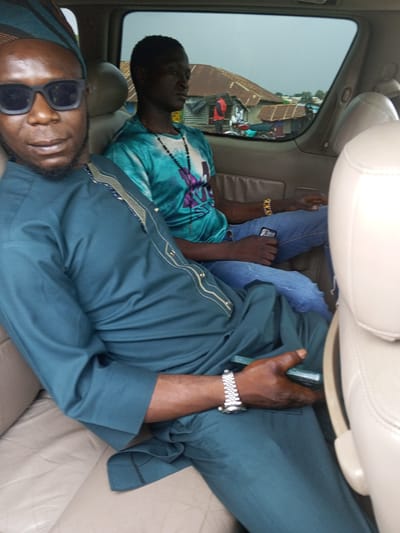 Two men are seated in the back of a vehicle. The man in the foreground is wearing a dark teal traditional outfit with gold trim and a matching head covering. He has on black sunglasses and a silver wristwatch. The man behind him is wearing a teal tie-dye t-shirt and blue jeans, and he is holding a dark-colored mobile phone. Outside the vehicle window, a glimpse of a settlement with corrugated metal roofs and some vegetation is visible, indicative of a scene in the city of Kerang, Nigeria. The interior of the vehicle appears to be upholstered in light-colored leather.