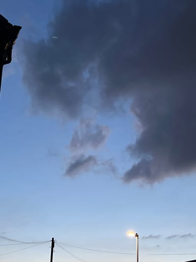 The image captures a twilight sky over Stoke-on-Trent, United Kingdom, with dramatic, dark clouds dominating the upper portion. A sliver of a building's roofline is visible in the upper left corner. A street lamp illuminates the lower right quadrant of the scene, casting a warm glow. Telephone poles and wires are present in the lower part of the image, suggesting an urban or suburban setting. The overall atmosphere is moody and atmospheric, indicative of impending or recent weather changes.