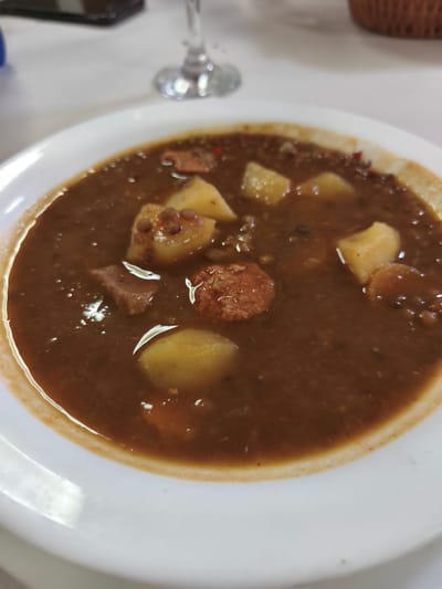 A close-up, slightly overhead shot captures a hearty bowl of dark brown stew, likely lentil stew (lentejas), served in a white, shallow dish. The stew is rich with ingredients, prominently featuring numerous chunks of light yellow potatoes, various pieces of meat (some dark brown, resembling beef or pork, and some reddish-brown, consistent with sliced chorizo or sausage), and small, dark lentils suspended in the thick broth. Small red specks, possibly from spices or a type of pepper, are also visible.
The white plate has a distinct, slightly raised rim around the edge of the stew. The background is softly blurred, showing parts of a clear stemmed wine glass on the left and what appears to be a woven basket or container on the right, suggesting an indoor dining setting, possibly a restaurant or home. The table surface beneath these blurred background elements is white. No people are visible, and there is no discernible text in the image. The dish appears warm and substantial, characteristic of comfort food. The style of the dish is consistent with traditional Spanish cuisine, aligning with the given location context of Polígon Industrial De Picassent, Spain.