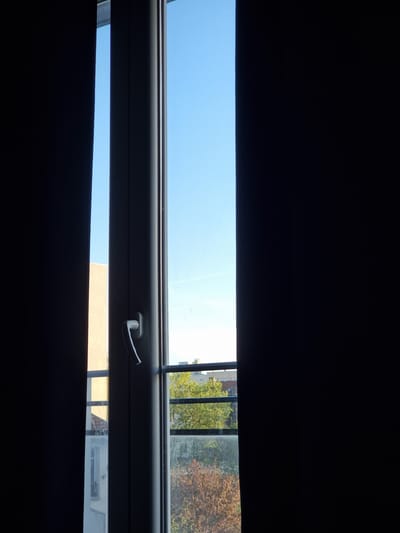 The media file presents a view from a darkened interior looking out through a partially opened window or blinds. The image is horizontally composed, with large, dark, undefined areas at the top and bottom, likely representing interior blinds or roller shades. A prominent horizontal strip across the middle of the frame reveals the exterior.

Through this opening, a clear, bright blue sky dominates the upper portion. Below the sky, the upper part of a window frame, light gray or white, is visible, extending across the frame. A white window handle is distinctly seen on the right side of this frame. Below the window frame, a narrower horizontal strip offers a glimpse of the outdoor environment.

This outdoor view includes lush green trees and trees with autumnal brown and orange foliage, suggesting a late summer or early autumn season. Parts of urban buildings are also visible; a light-colored (possibly beige or pale orange) building facade appears directly outside, and other indistinct structures are present in the distance on the right. A dark vertical element, possibly a balcony railing or another part of the window structure, is also discernable on the right side.

The bright daylight outside, contrasting with the dark interior, indicates a daytime setting, likely with clear weather. There are no people visible, and no discernible text is present in the image. The scene suggests a vantage point from a building in an urban or suburban area, consistent with the given location context of Clichy, France.