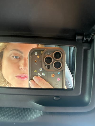 In a car's sun visor mirror, a woman's reflection is visible. She has blonde hair and is wearing makeup, with glossy pink lipstick and blue eyeshadow. Her left eye is looking directly at the camera, while her right eye is obscured by the phone she is holding up to take the picture. The phone has a floral patterned case with small daisies and sunflowers. The woman is wearing a necklace. The background is a blurred car interior, with a hint of a leather seat. The mirror is framed by a black sun visor, and a small button is visible below the mirror. The overall lighting suggests it is daytime, possibly sunny, with light reflecting on the woman's face and the mirror.