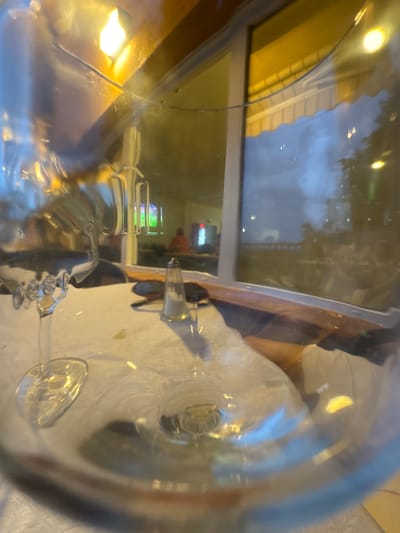 This image, taken in El Escorial, Spain, provides a view through a glass, likely a wine glass, set on a table covered with a white tablecloth. The glass itself is a prominent element, reflecting its surroundings.
In the background, visible through the glass and a window, is an indoor scene. There appears to be a television screen displaying a green and red image, possibly a game or broadcast. A person wearing an orange shirt is visible sitting at a table in this indoor area. The window reflects an outdoor scene with a cloudy sky and some trees, illuminated by warm, artificial lighting from an awning or building structure.
On the table, in front of the glass, are a salt shaker and a pepper shaker, typical of a restaurant setting. The lighting in the scene is a mix of ambient indoor light and reflections from outdoor elements. The overall impression is that of a dining establishment, possibly a restaurant or cafe, in El Escorial.