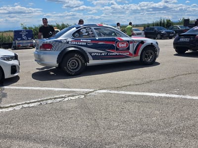 In Akatsievo, Bulgaria, on a bright, cloudy day, a racing car is parked on an asphalt surface marked with white lines. The car, predominantly silver with blue and red racing stripes, is emblazoned with various sponsor logos, including "Unil Lubricants" and "Opaljet Competition." A man with short, dark hair and sunglasses is standing next to the car, appearing to be involved in some activity related to it. In the background, a blue trailer with "VIDA RACING TEAM" graphics and a Bulgarian license plate is visible, as well as other vehicles and a line of trees bordering a distant field. The overall scene suggests a racing event or preparation.
