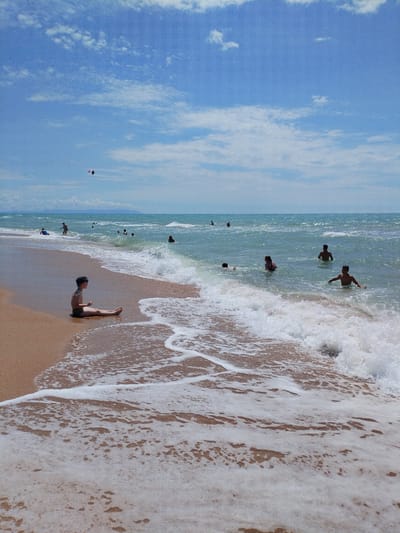 A wide shot captures a beach scene with the sea, sand, and sky. In the foreground, a young boy sits on the wet sand near the water's edge, wearing swim trunks and goggles. He is facing left and appears to be looking out at the sea. Gentle waves are washing over the sand, creating white foam.
Further out in the turquoise water, several people are swimming and wading. A kite can be seen flying in the blue sky with scattered white clouds. The background shows a hazy coastline. The overall atmosphere is that of a sunny day at the beach. The location is identified as Vinogradny, Russia.