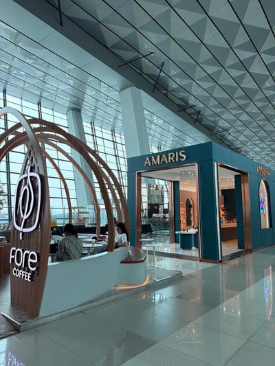 The image depicts an interior view of a modern airport terminal, specifically featuring a "fore Coffee" establishment and an "AMARIS" store. The "fore Coffee" area has a distinctive arched wooden structure with a glowing logo. Two individuals are seated at a table within this coffee area, one looking at their phone and the other appearing to be engaged in conversation. The "AMARIS" store is a sleek, teal-colored retail space with gold lettering and a prominent star motif on its facade. The environment is characterized by a high, geometrically patterned ceiling and large glass windows that allow ample natural light. The polished floor reflects the interior lighting and structures. The location is identified as Rawa Burung, Indonesia, suggesting this is an airport within that region.