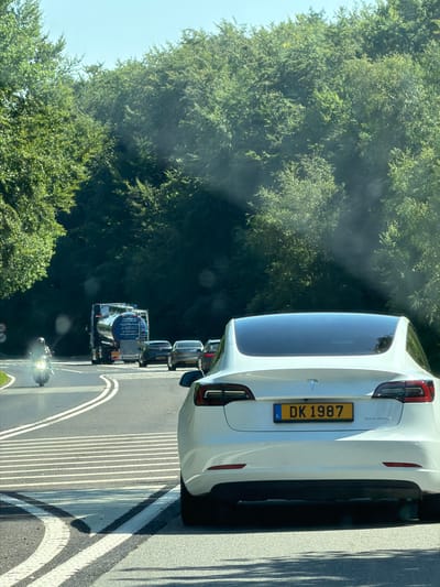 In Niederanven, Luxembourg, on a sunny day, a white Tesla Model 3, license plate DK 1987, is seen from behind, positioned on a road with white lane markings and a crosswalk. The car is in the foreground, partially obscuring the view of other vehicles. Ahead of the Tesla, a large blue tanker truck is visible, followed by other cars. To the left, a motorcycle rider is traveling on the road, and the surrounding environment is characterized by a dense green forest on both sides of the road and a clear blue sky.